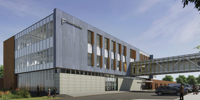 Modern three-story building with a sleek design featuring large windows, metal paneling, and wooden accents. A glass-enclosed skybridge connects to another structure. The building is surrounded by greenery, with people walking and cars parked in the foreground.