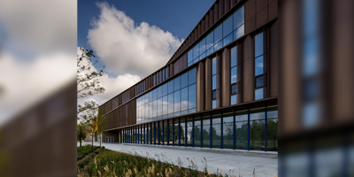 The image shows a modern building with a sleek, brown exterior and extensive glass windows reflecting the sky. In the foreground, there is a landscaped area with plantings alongside a paved pathway, contributing to the contemporary aesthetic of the structure.