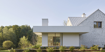 A contemporary white house featuring a flat roof and large windows, set amidst green trees and landscaped grass. The design blends modern architecture with natural surroundings, creating a peaceful atmosphere.