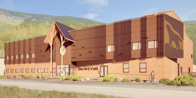 A contemporary wooden building stands in the foreground, set against a backdrop of mountains. The structure features an artistic representation of a wolf on its side, gabled roof with a central circular element, and several large windows. A pathway leads up to the entrance, where a few people are seen walking.