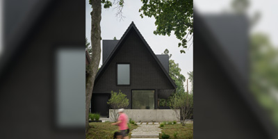 Modern house with a steep dark gabled roof, large windows, and a minimalist design surrounded by greenery, featuring a concrete base and a pathway leading to the entrance.