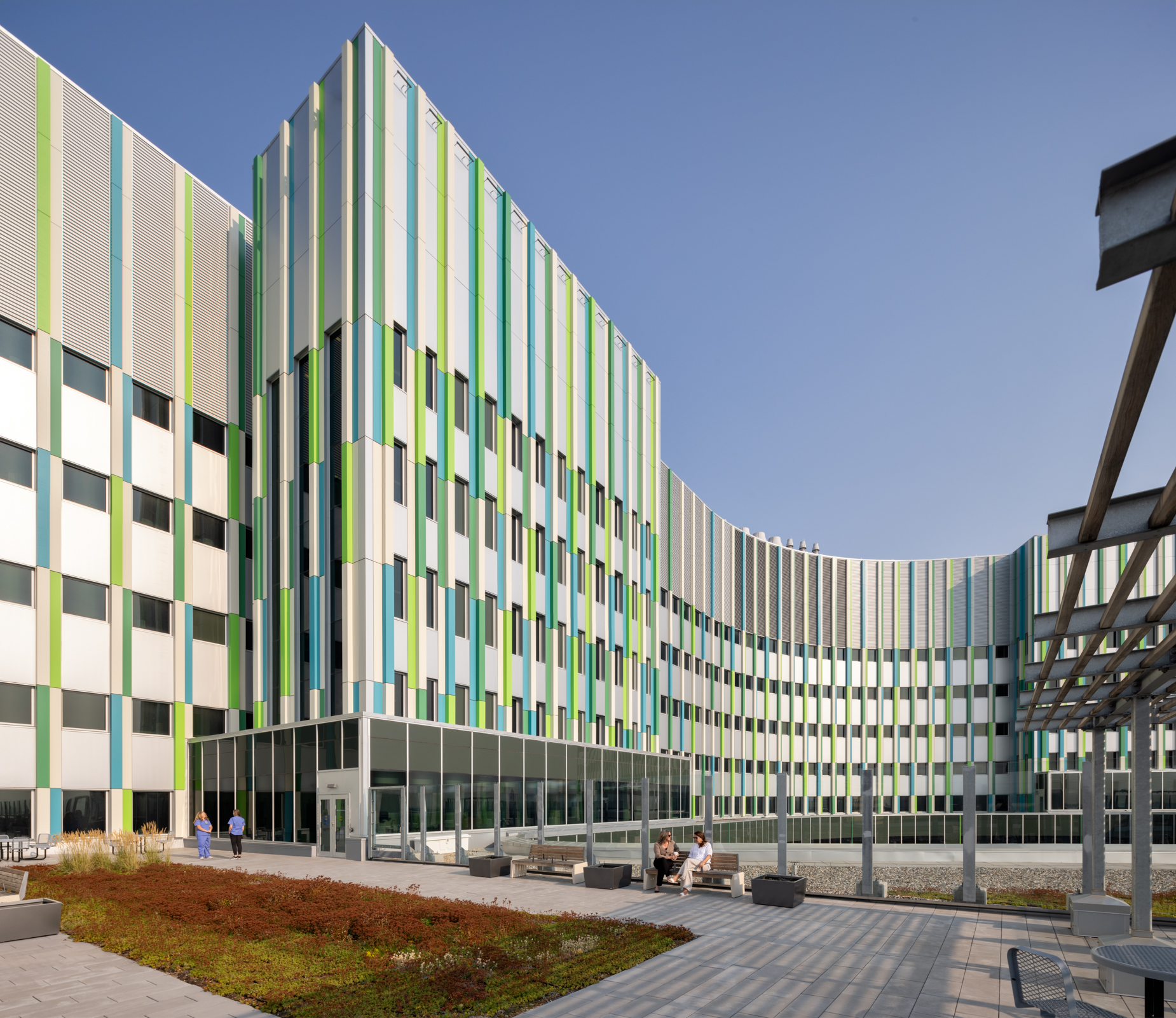 Modern building with green and white striped facade, surrounded by a landscaped courtyard and people enjoying the outdoor space.