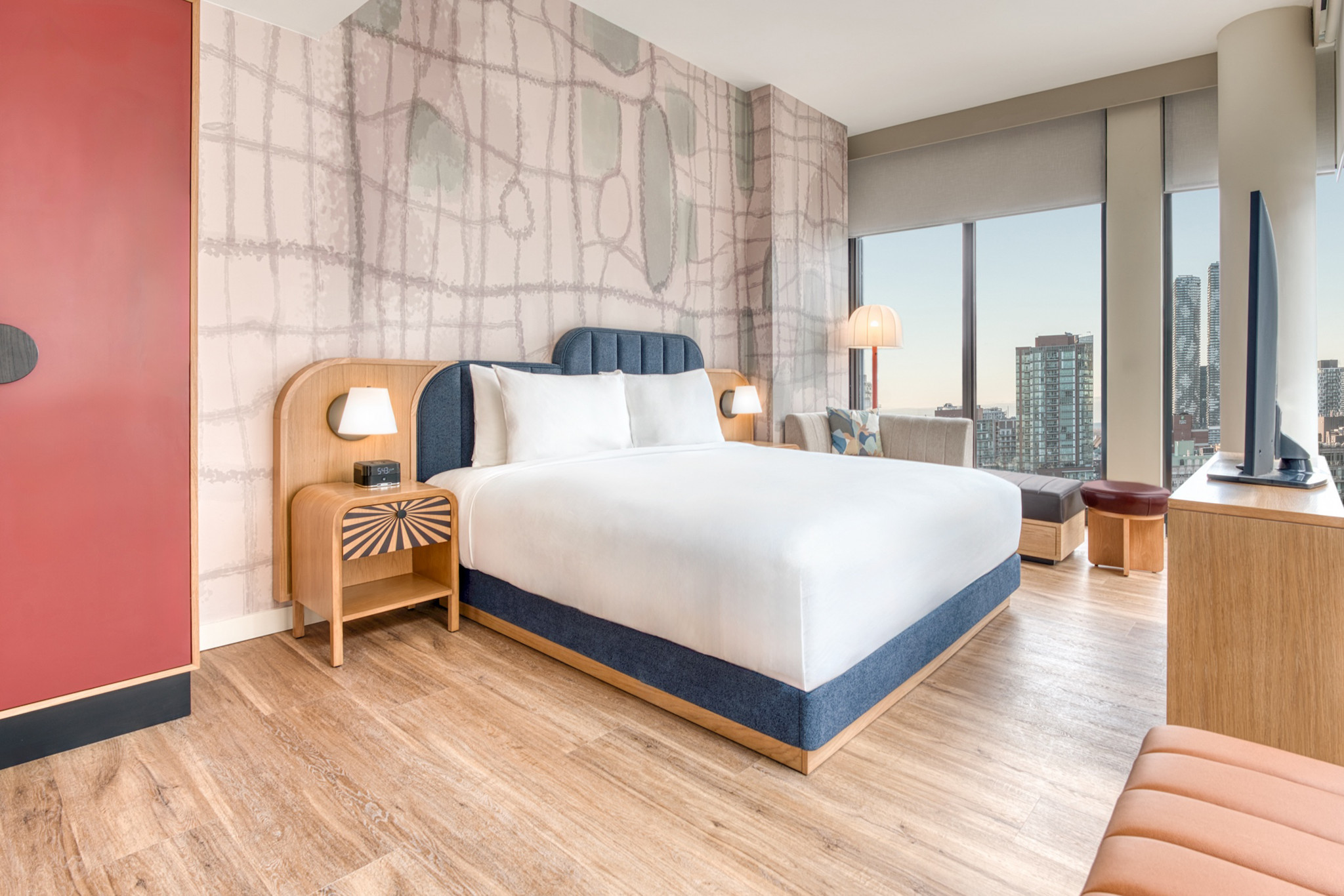 Modern hotel room with wood flooring, abstract wall art, a platform bed with blue accents, and large windows offering a city skyline view.