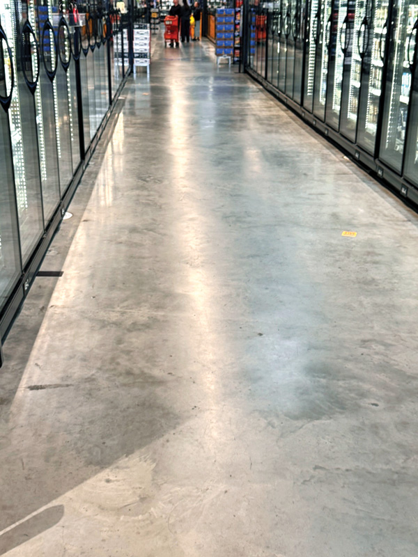 A spacious grocery store aisle lined with glass-fronted refrigerators, leading to shoppers in the distance. Smooth concrete floor visible.