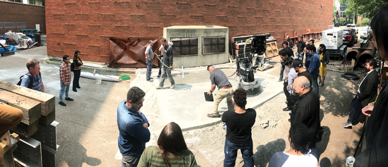 A group observes a construction demonstration in a bustling outdoor area, surrounded by building materials and machinery.
