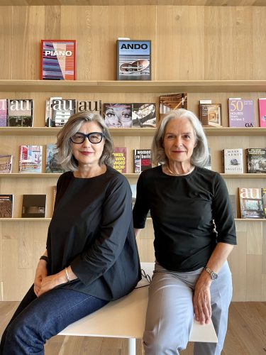 Two women with gray hair sit side by side on a white bench in front of a wooden shelf filled with various architecture magazines.