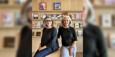 Two women with gray hair sit side by side on a white bench in front of a wooden shelf filled with various architecture magazines.