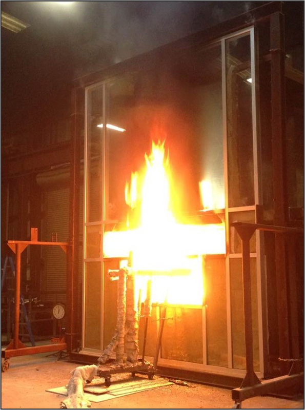 Intense flames erupt from a window frame in a lab setting, surrounded by smoke and metal structures, indicating a controlled fire test.