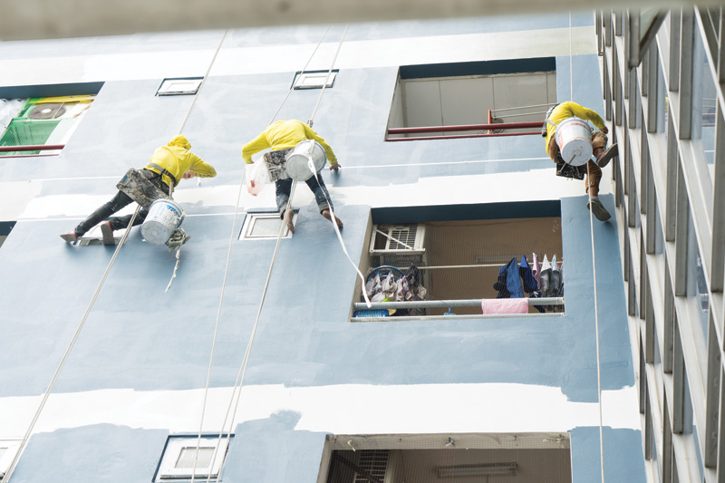 Three workers in yellow jackets rappel down a building, painting the exterior wall with blue paint while a window shows laundry inside.