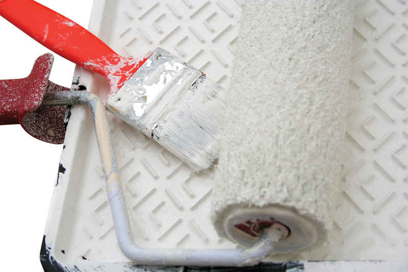 A paint roller and brush with a red handle sit in a paint tray, covered with white paint, on a textured white surface.