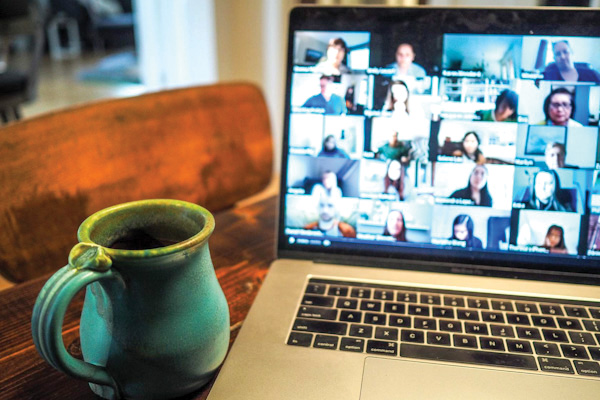 A laptop displays a video conference with multiple participants, next to a rustic blue mug filled with coffee on a wooden table.