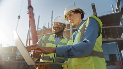 Two construction workers in safety vests and helmets review plans on a laptop, with a building site in the background.
