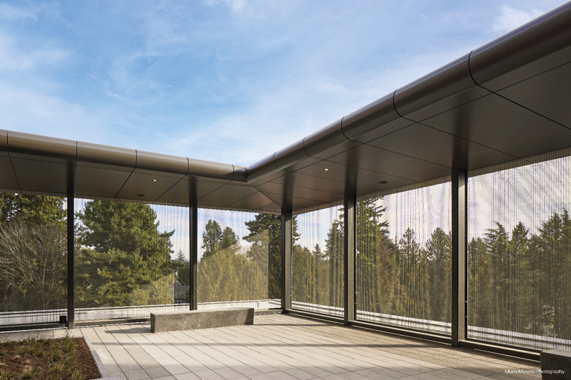 Modern outdoor space with mesh screens and a concrete bench, surrounded by greenery under a clear blue sky.