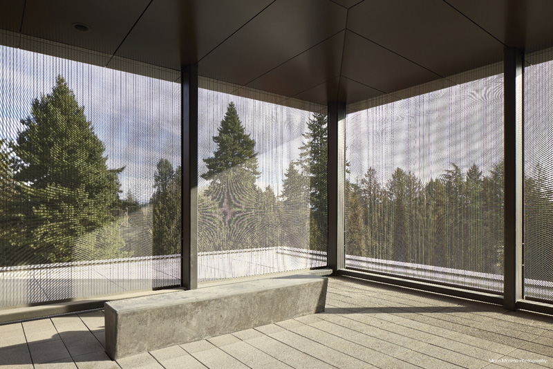 A minimalist interior space featuring a concrete bench, surrounded by mesh walls displaying views of lush green trees and a serene sky.