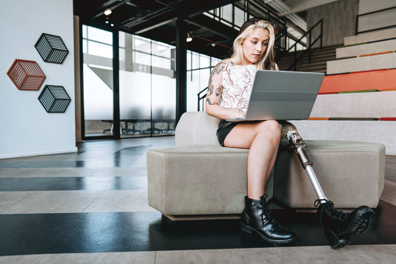 A person with a prosthetic leg sits on a couch, working intently on a laptop in a modern, bright workspace.