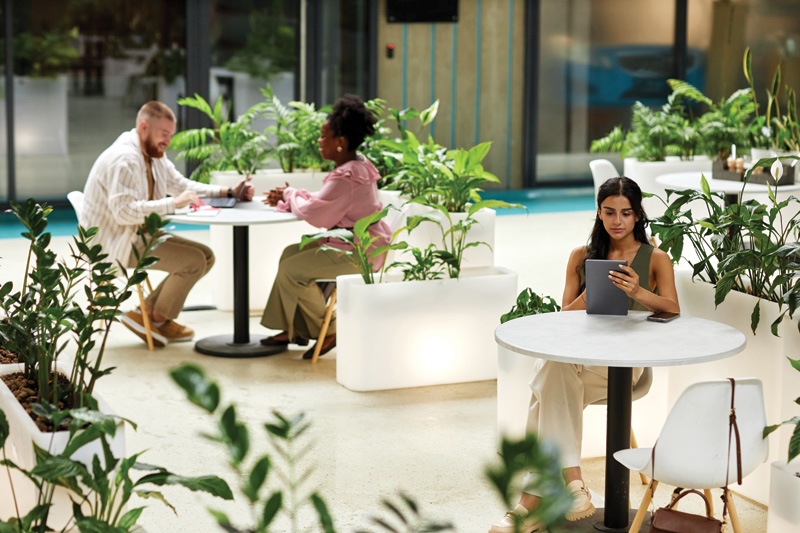 A modern cafe scene with two individuals engaged in conversation at a table surrounded by lush greenery, while another sits on a device.
