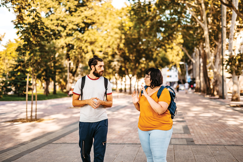 Two people walk along a sunlit park pathway, engaged in conversation amidst lush trees and a serene outdoor setting.