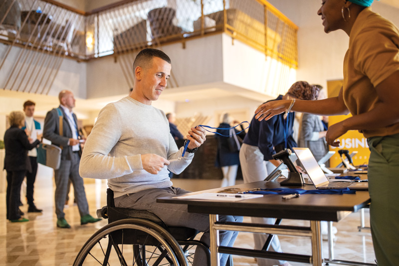 A person in a wheelchair interacts with an event staff member at a registration desk in a bustling lobby filled with attendees.