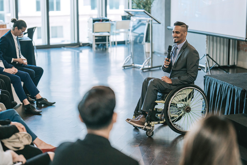 A speaker in a suit sits in a wheelchair, addressing an audience in a modern conference room with large windows.