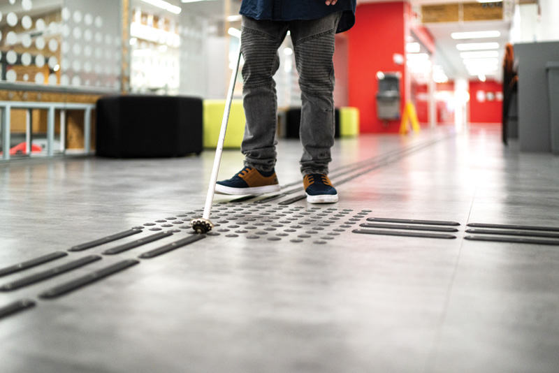 A person using a white cane navigates a floor featuring tactile paving for accessibility in a modern indoor space.