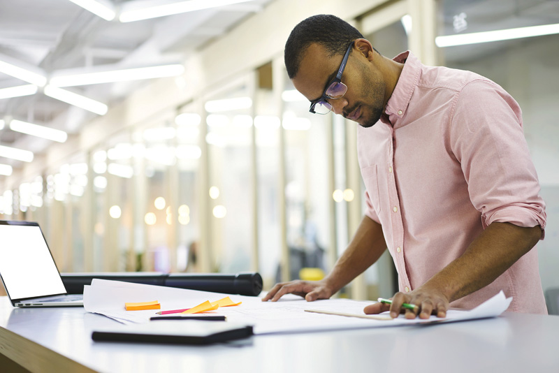 A person in a pink shirt reviews blueprints and sketches on a table, with a laptop and colorful stationery nearby in a modern workspace.