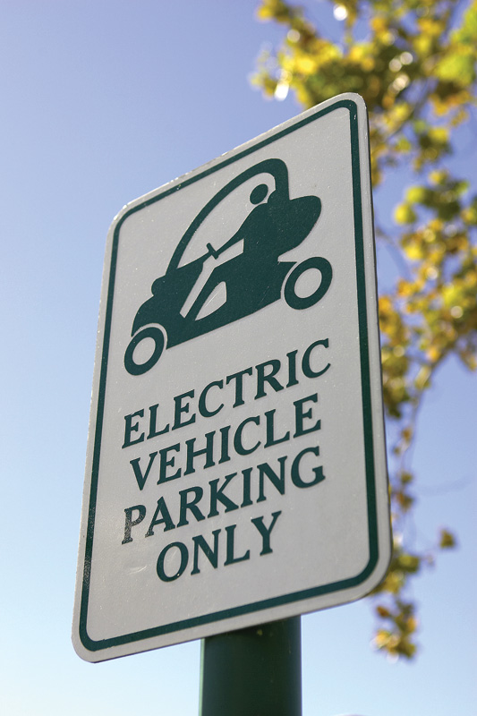 Sign indicating designated parking for electric vehicles, featuring a graphic of an electric car, with a clear blue sky in the background.