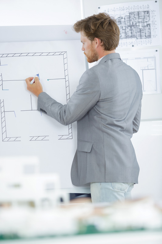 A man in a gray blazer draws a floor plan on a whiteboard, surrounded by architectural blueprints and models.