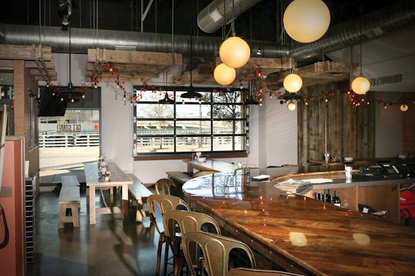 Modern restaurant interior with wooden bar, metal chairs, and large windows, illuminated by round pendant lights and cozy decor.
