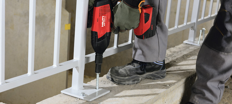 A worker in gloves uses a power tool to secure a metal railing to a concrete surface. Safety is emphasized with protective footwear.