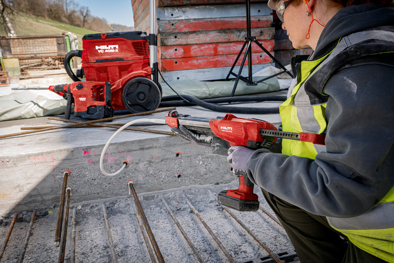 A construction worker uses a Hilti tool on a concrete surface with exposed steel rebar, and a Hilti vacuum is visible in the background.