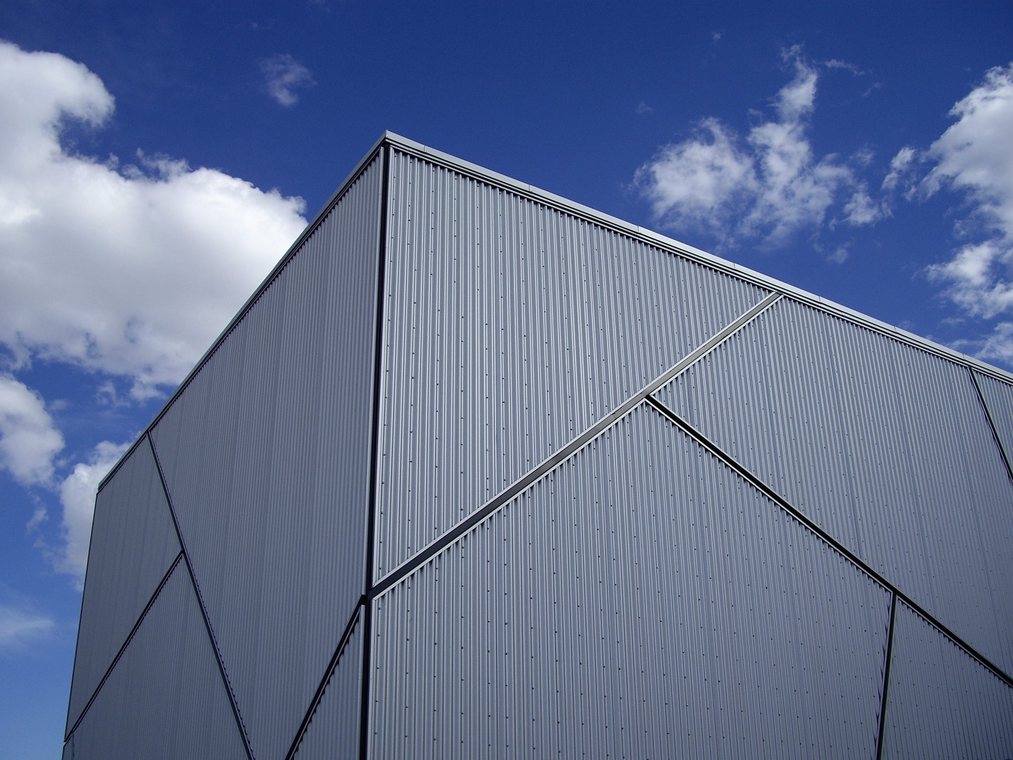 Modern building corner with sleek metal facade, framed against a bright blue sky with scattered clouds.