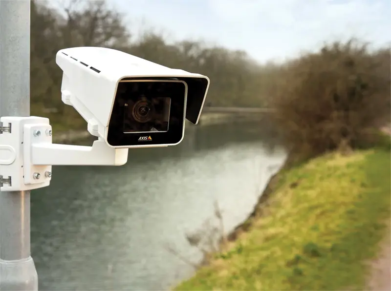 A white outdoor security camera mounted on a pole overlooking a serene riverbank surrounded by trees and greenery.