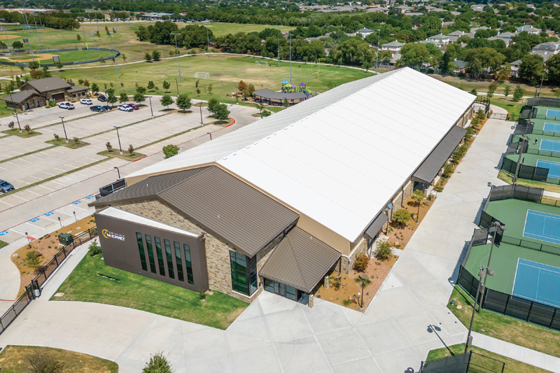 Aerial view of a modern sports facility with a large white roof, surrounded by greenery, tennis courts, and a park area.