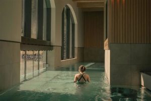 A woman in a swimsuit wades through a serene, partially submerged pool in a modern, light-filled spa with large arched windows.