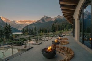 Modern outdoor lounge with fire pits, overlooking a serene lake and majestic mountains at sunset.