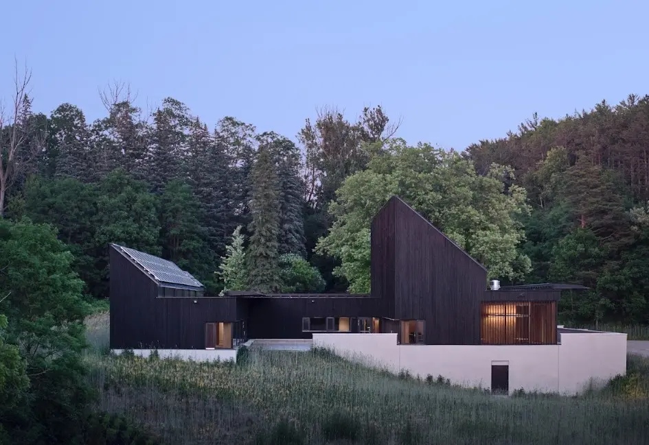 Modern, multi-level house with dark wooden exterior, set in a lush green landscape, surrounded by trees and gentle hills at dusk.