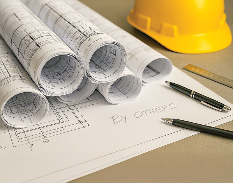 Blueprints rolled up on a workspace, with a yellow hard hat and pens nearby, displaying the note "By Others."