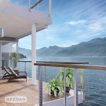 A serene balcony overlooking a calm lake and mountains, featuring wooden flooring, plants, and a lounge chair under a clear sky.