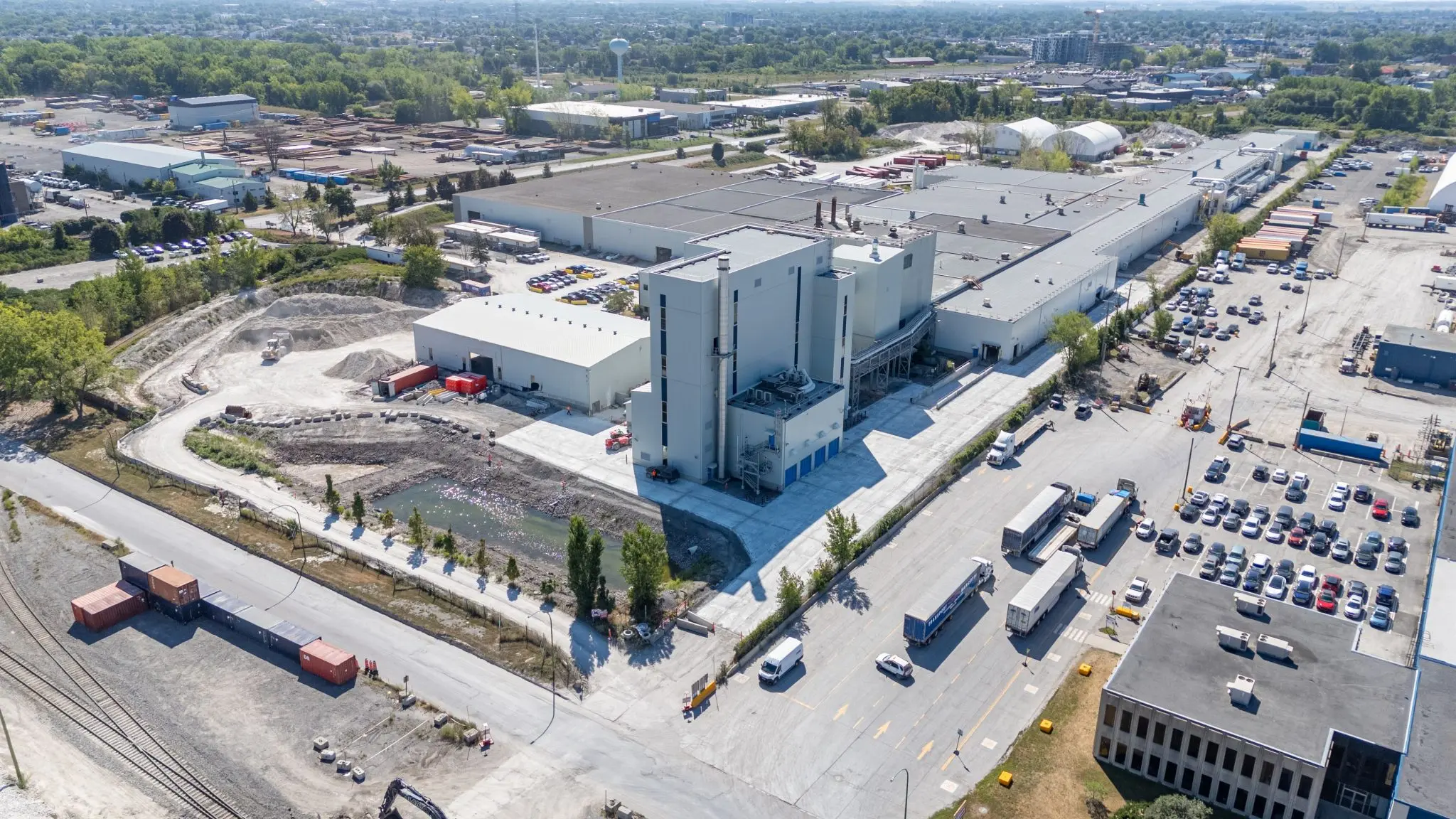 Aerial view of a large industrial facility with trucks, parking lots, and nearby construction areas amidst green landscaping.