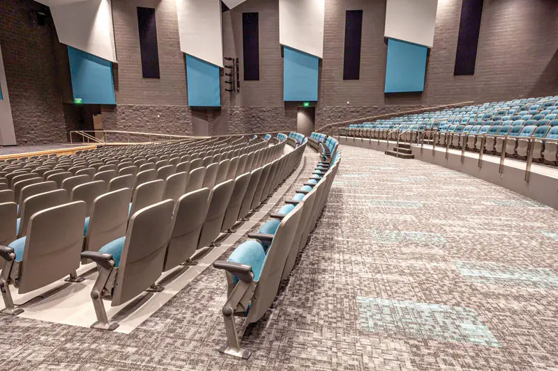 A modern auditorium featuring curved rows of gray seats with blue accents, set against textured gray walls and vibrant blue panels.
