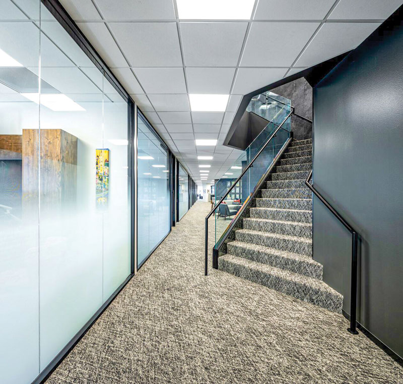 Modern office corridor featuring glass walls, carpeted flooring, and a staircase with a glass railing.