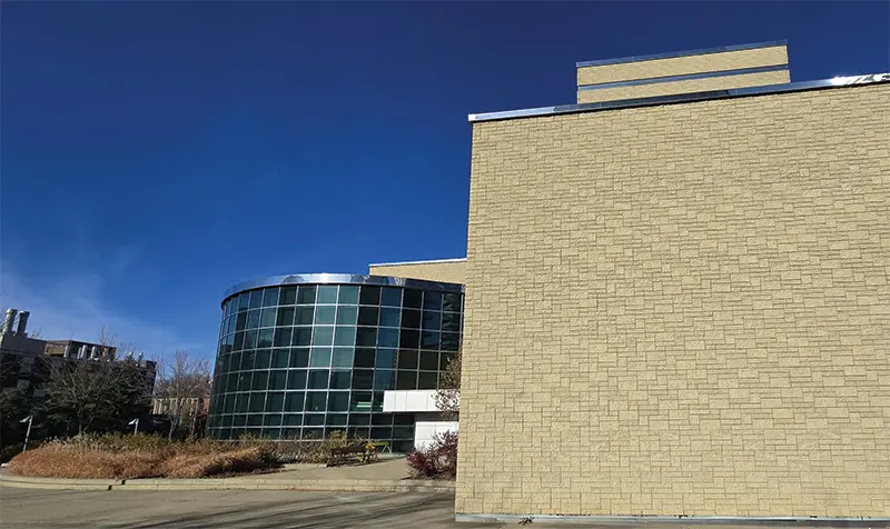 Modern building with a large glass facade and textured beige wall, set against a clear blue sky. Sparse landscaping in the foreground.