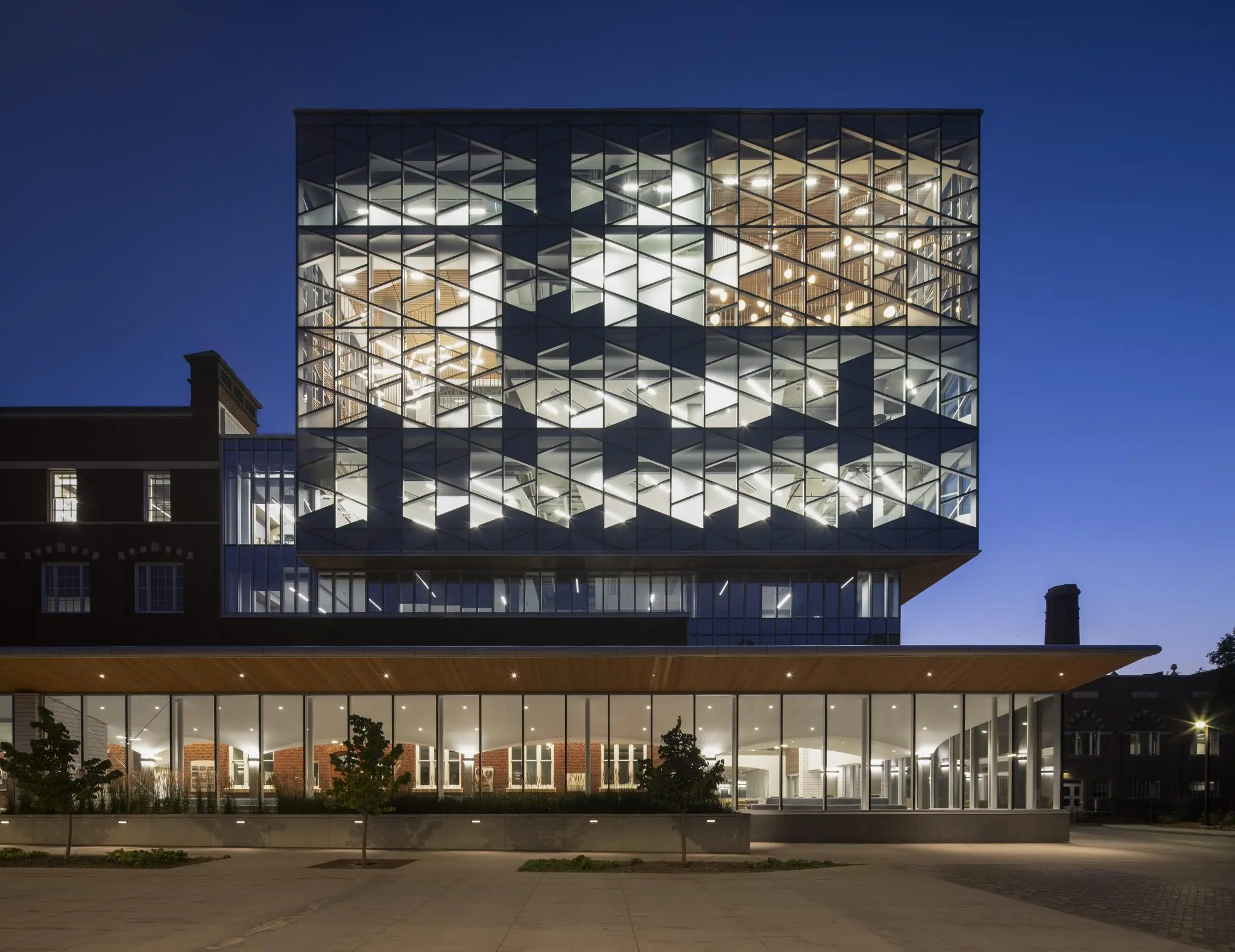 Modern multilevel building with a glass façade, illuminated at dusk, adjacent to a classic brick structure. Greenery in the foreground.