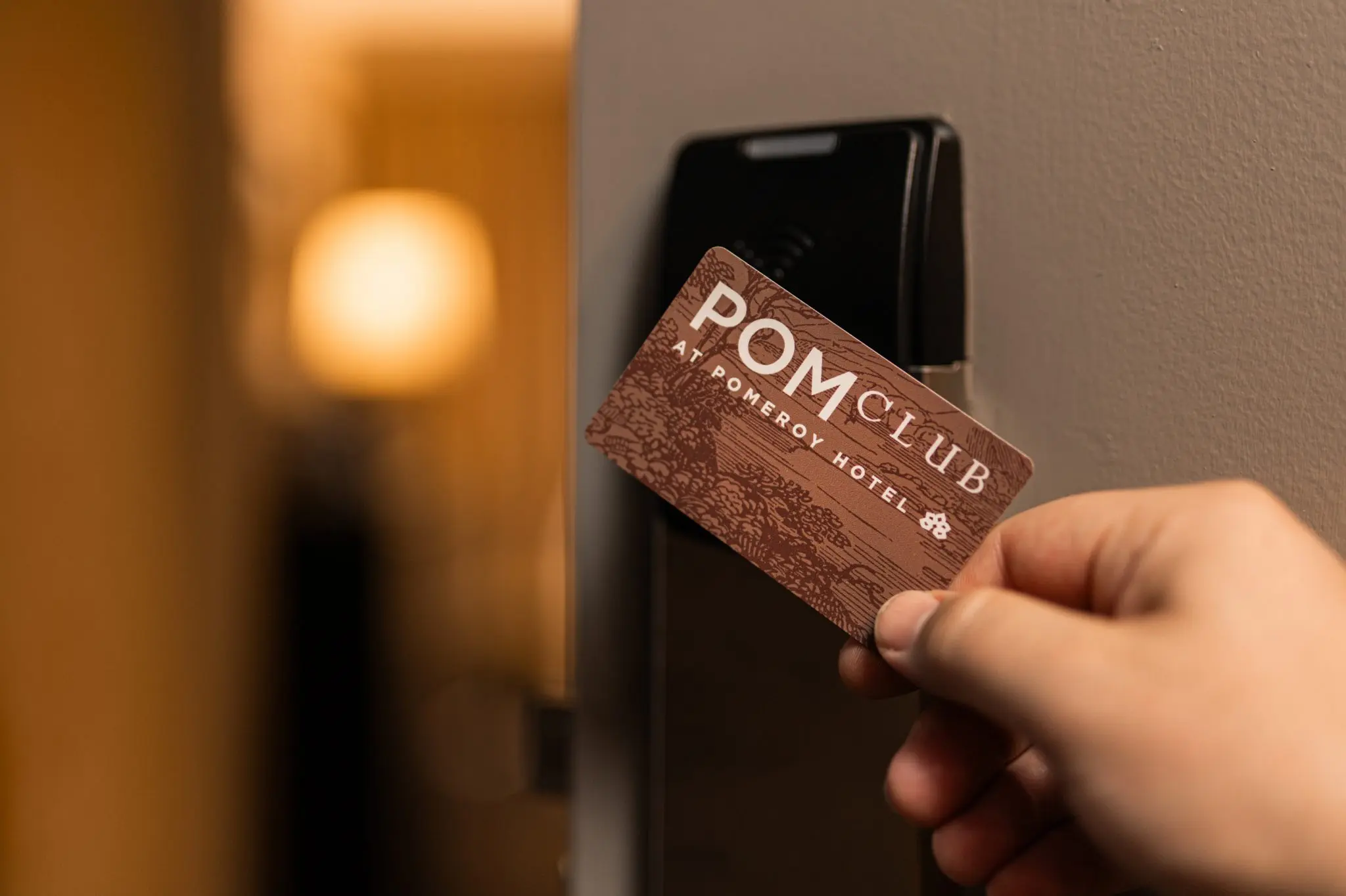 A hand holds a POM Club hotel card near an electronic door lock, with a softly lit background.