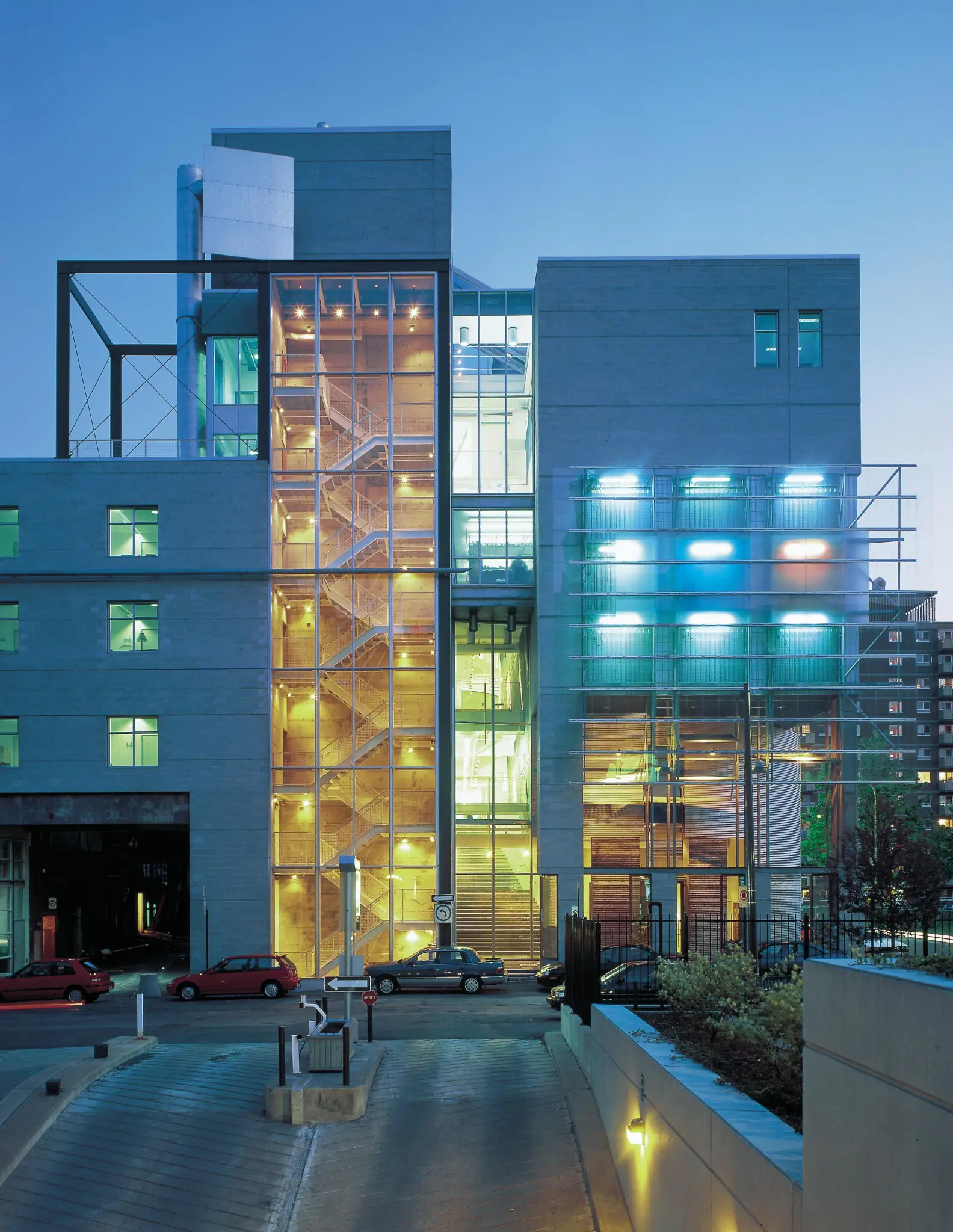 Modern building with glass-encased stairwell lit warmly at dusk, adjacent to a multi-story structure with blue neon lights.