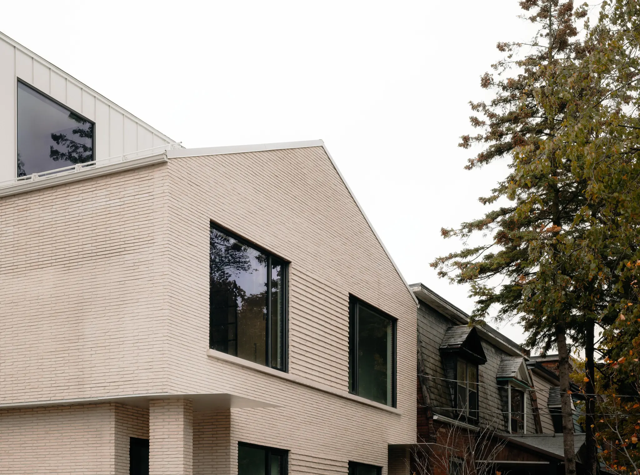 Modern house with light brick facade, large black-framed windows, and a sloping roof. A tree with autumn leaves stands to the right, under a cloudy sky.