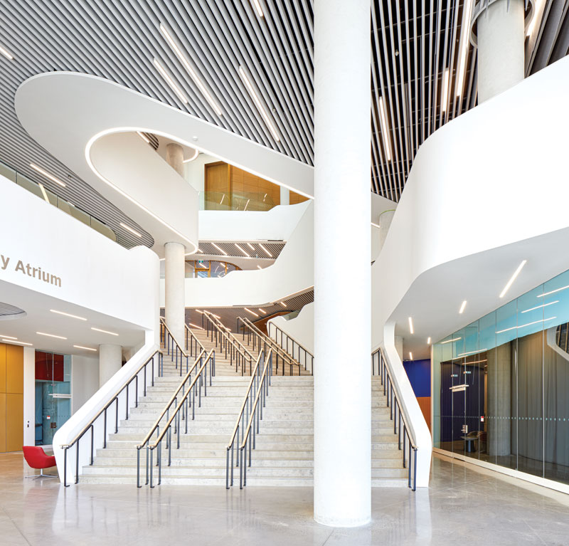 Modern spacious atrium with wide staircases, sleek white curved railings, glass walls, and a ceiling with linear black and white panels.