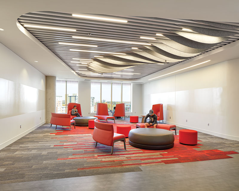Modern lounge area with red seating, large windows, patterned carpet, wavy ceiling design, and two people using electronic devices.