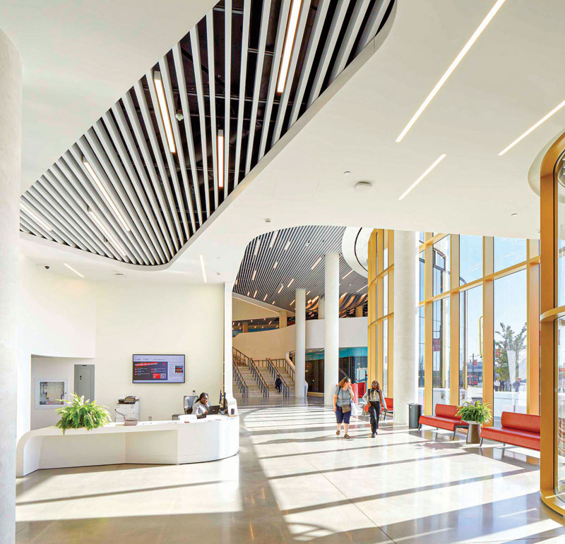 Modern spacious lobby with high ceilings, large windows, white pillars, red seating, and people walking near a white reception desk.