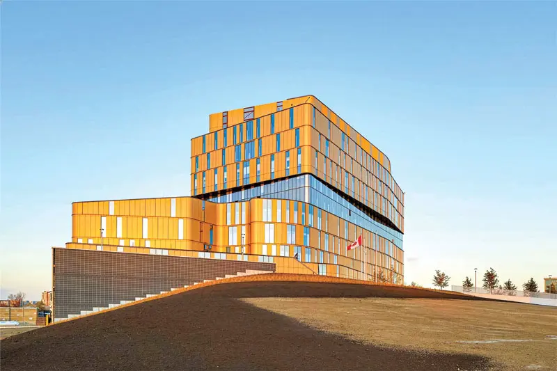 Modern, multi-story building with orange and blue panels under a clear blue sky, situated behind a gently sloping dirt hill.